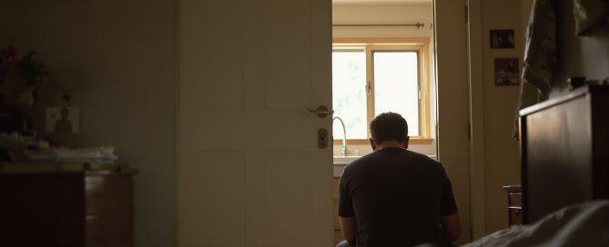 Man sitting alone on the edge of a bed in a dimly lit room seen through a doorway