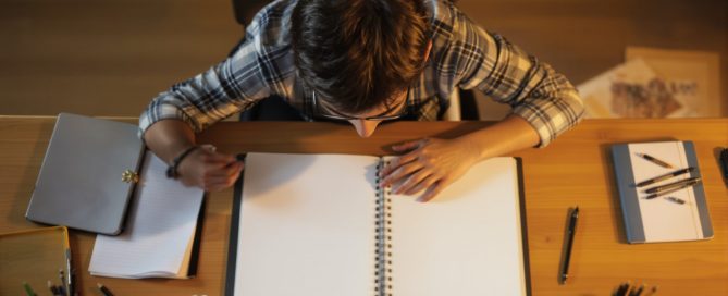 Frustrated young man hunched over a blank notebook surrounded by crumpled paper at a desk